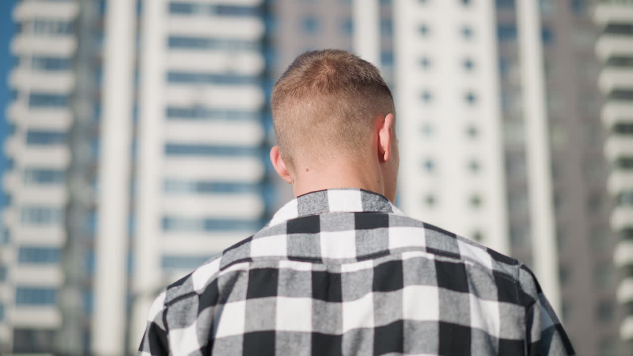 Back view of young man in black and white checkered shirt walking outdoors under bright sun during clear day with tall modern building blurred in background and soft shadows