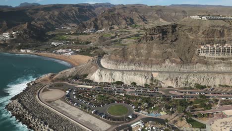 Slow-aerial-forward-flight-over-parking-cars,-road-around-coastline-and-Playa-de-Amadores-in-background,Gran-Canaria