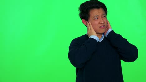 젊은 아시아인 남자가 큰 소리에 대해 귀를 감고 미소를 지었습니다. - 그린 스크린 스튜디오