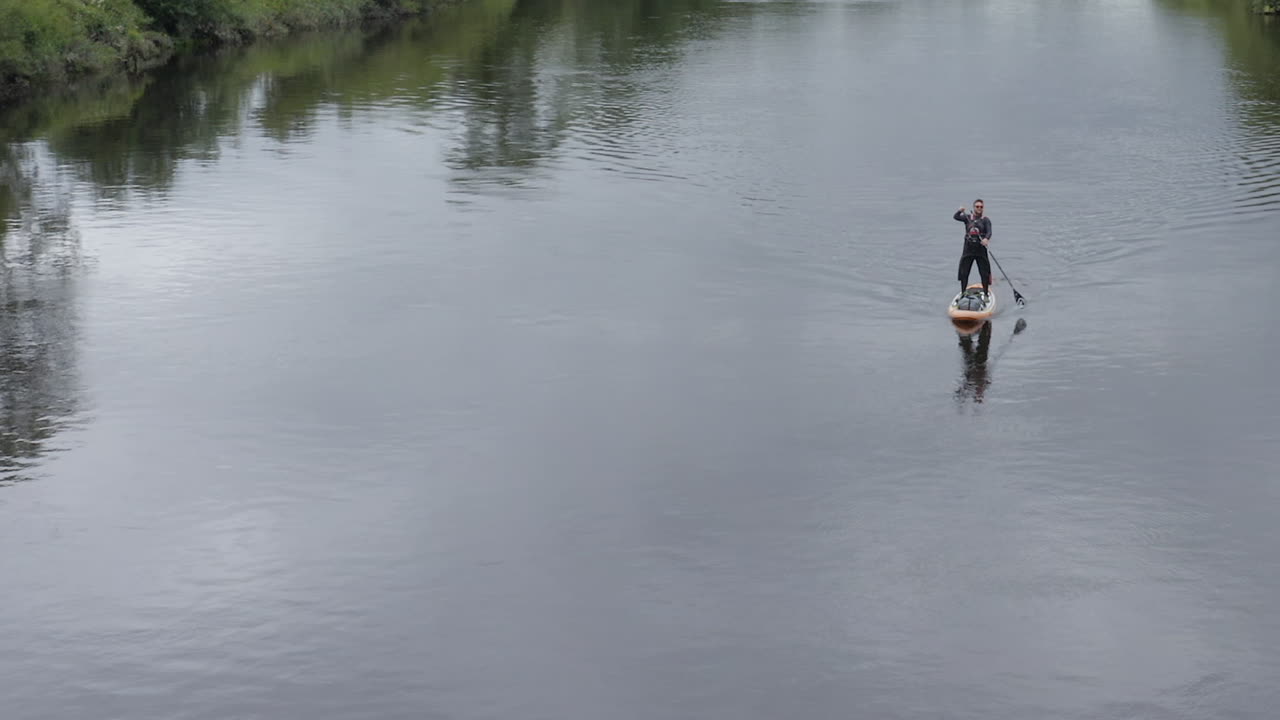 Caucasian man in sunglasses paddles paddleboard on calm rural river