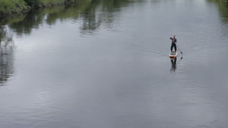 caucasian man in sunglasses paddles paddleboard on calm rural river