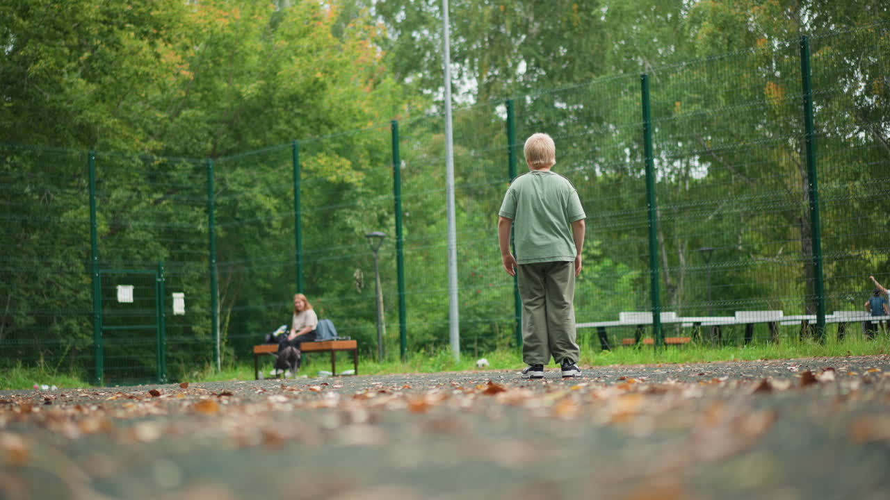 White Boy Practicing Recall Command With Dog On Court, Focused Posture And Hand Gesture, Attentive Pet Approaching For Reward, Green Fenced Park Backdrop With Bench Spectator, Patient Training Session