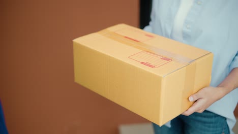 close up parcel in hand of blue uniform delivery man to customer front of the house from shopping online with good service. courier man delivering a cardboard box postal package to destination.