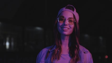 vista cercana de una adolescente caucásica con gafas y gorra sonriendo a la cámara al aire libre por la noche