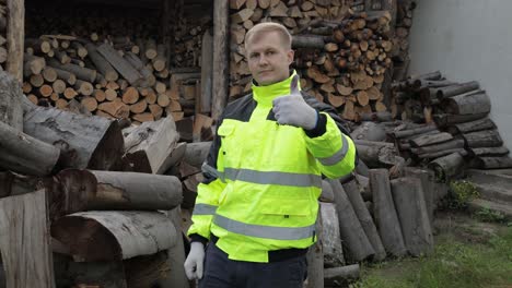 Lumberjack-in-reflective-jacket.-Man-woodcutter-show-thumbs-up.-Sawn-logs,-firewood-background