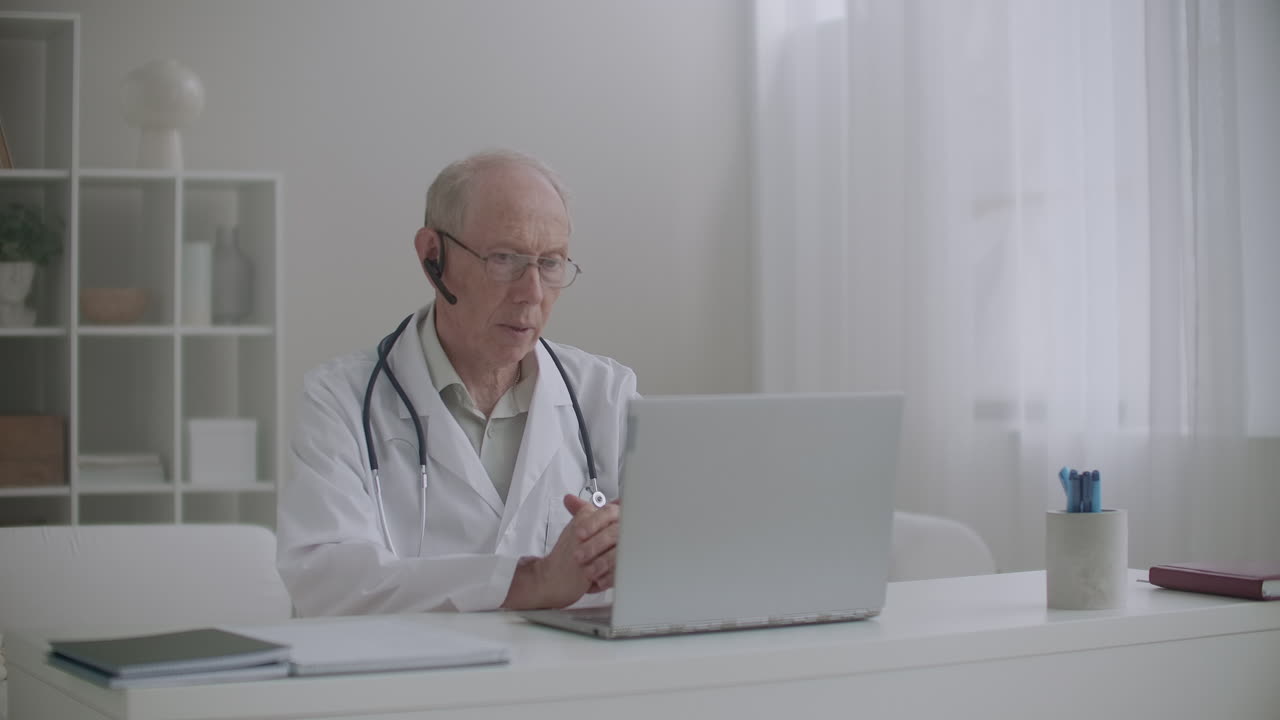 un médico anciano está consultando al paciente en línea sentado en la oficina de la clínica y comunicándose por internet en una computadora portátil