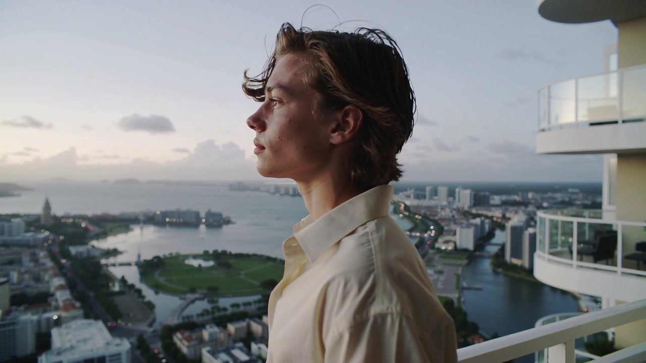 Camera moving in, man standing on balcony railing gazing at bay, thinking in buttonup shirt