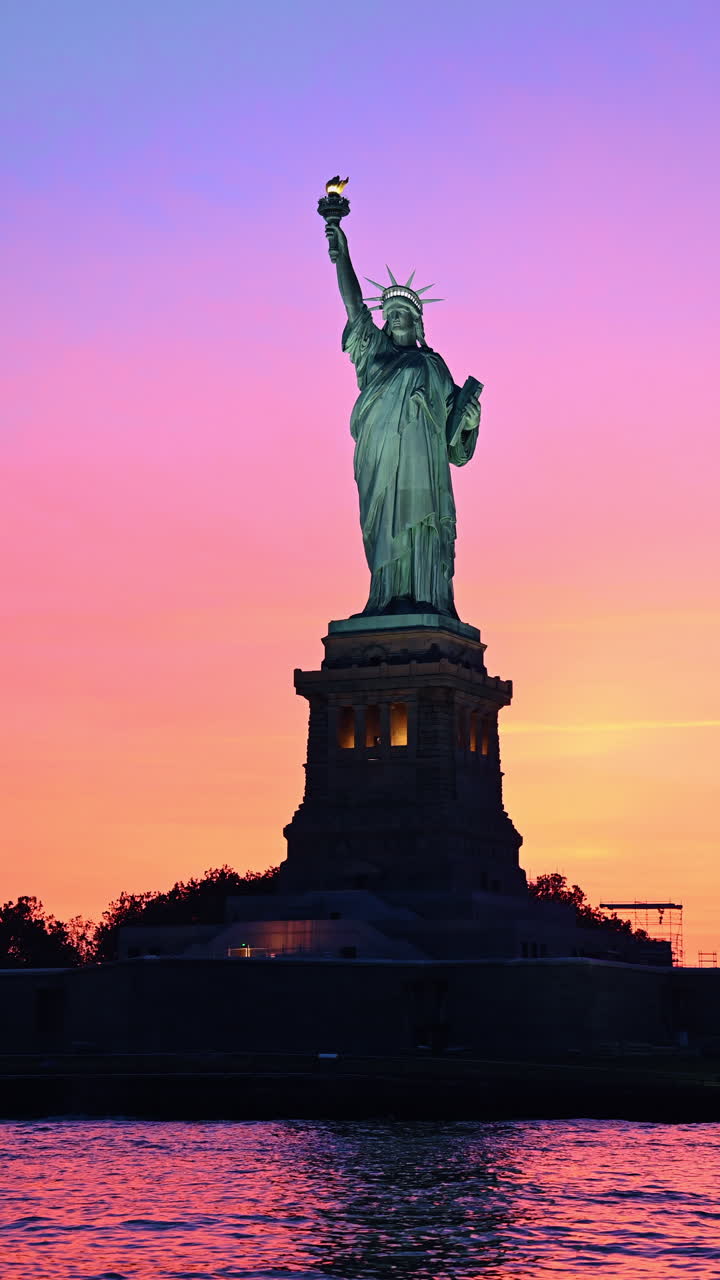 New York, AS, 9 Agustus 2025: Siluet Patung Liberty dengan latar langit matahari terbenam merah muda cerah NYC. Bidikan vertikal Patung Liberty di Pelabuhan New York saat langit matahari terbenam berwarna merah muda dan oranye yang penuh warna.