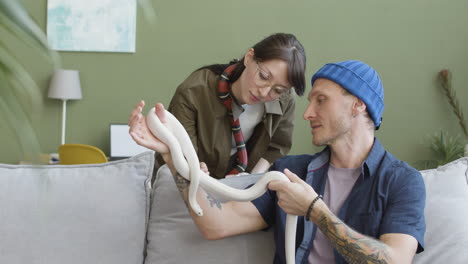 Couple-Holding-Pet-Snakes-And-Talking-Together-While-The-Man-Is-Sitting-And-Woman-Is-Leaning-On-A-Comfortable-Sofa-At-Home