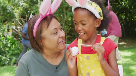 animation of happy african american grandmother and granddaughter painting easter eggs in garden