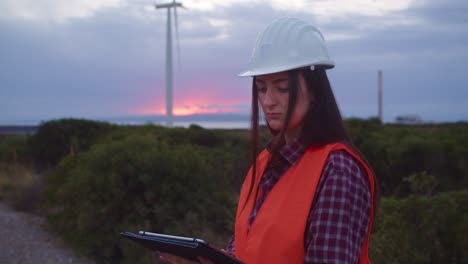 woman engineer checks tablet to inspect wind turbines parameters, slomo, sunset