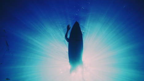 Underwater-silhouette-view-of-surfer-paddling-on-board,-light-rays-surround-shadow