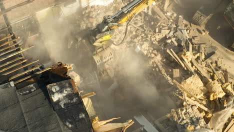 aerial view of a construction site where an excavator's arm is demolishing a building, creating dust clouds