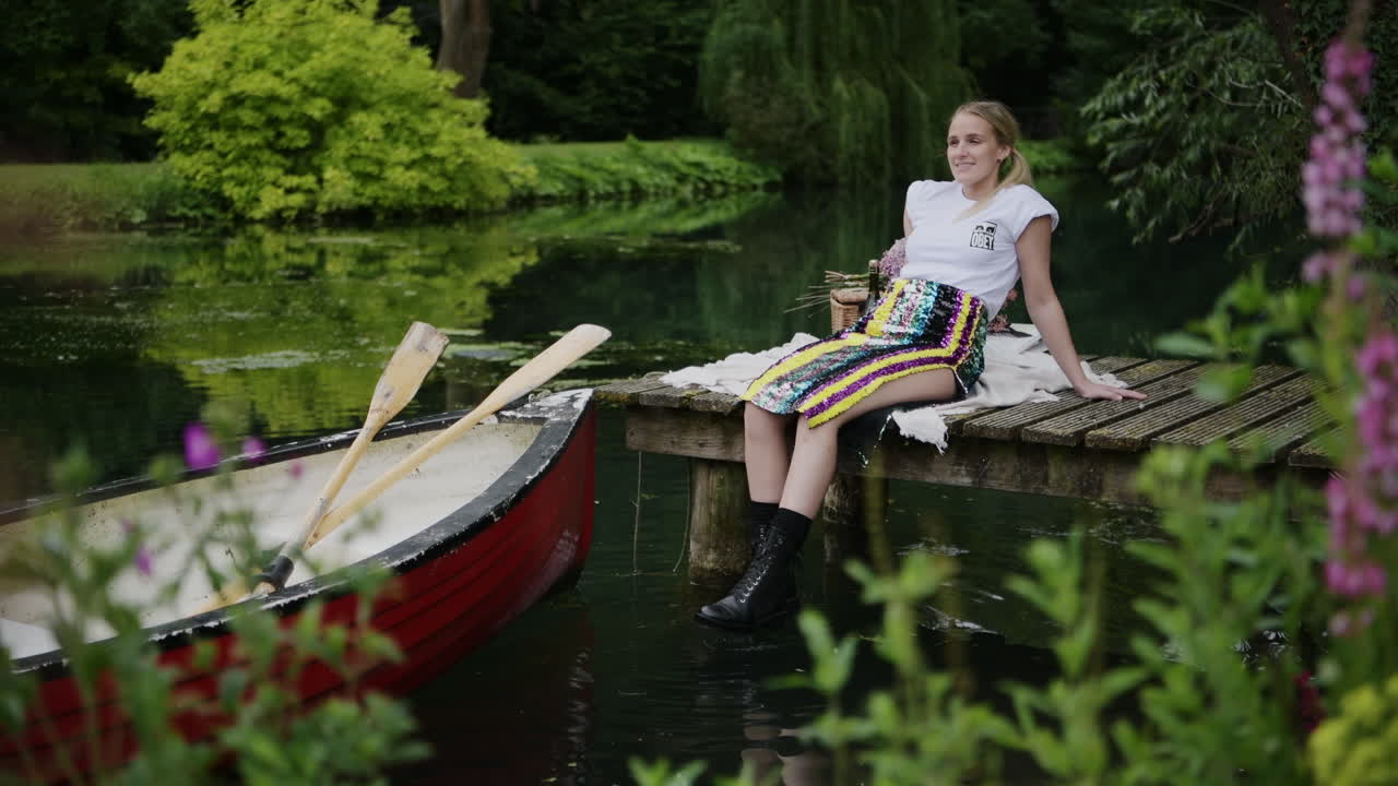 A Beautiful Fashion model Sitting By The Lake With A Boat Floating in an editorial photoshoot - full shot