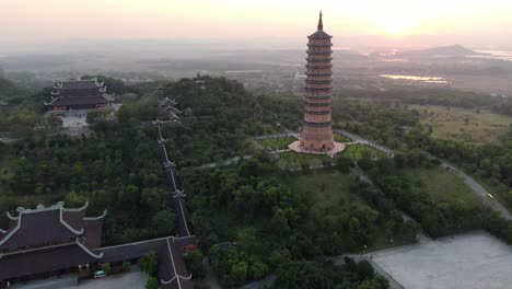 drone aerial view in vietnam flying over a buddhist temple area and pagoda filled with green trees in front of the sun in ninh binh at sunset