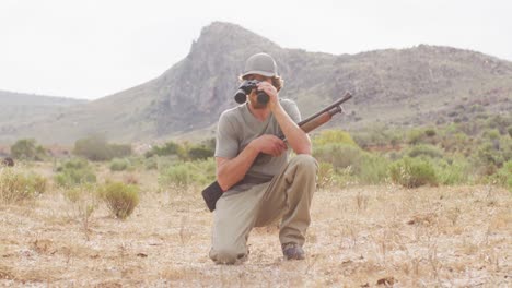 kneeling caucasian male survivalist holding hunting rifle, looking using binoculars in wilderness