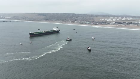 Toma-Aérea-De-Paralaje-Del-Mar-En-La-Costa-De-La-Playa-Pucusana-En-El-Océano-Pacífico-Con-Botes-Flotantes-Aislados-Y-Un-Gran-Carguero-Con-Vistas-A-La-Playa-Y-Los-Edificios