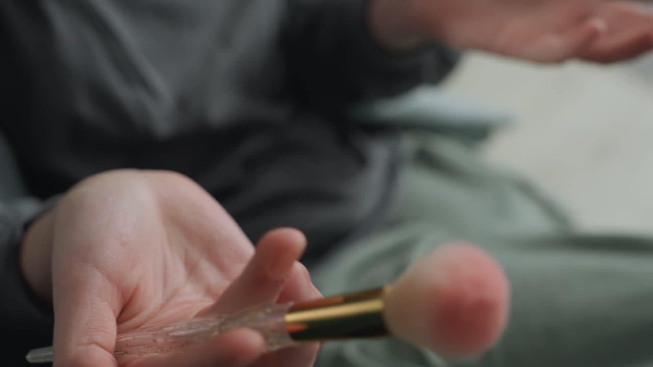 Mani in primo piano che tengono un pennello da trucco rosa, dettaglio di pizzo delicato, creatrice di bellezza che testa le setole, movimento tattile lento, toni pastello tenui, sfondo verde tenue, attenzione sui polpastrelli e sulla texture del pennello.