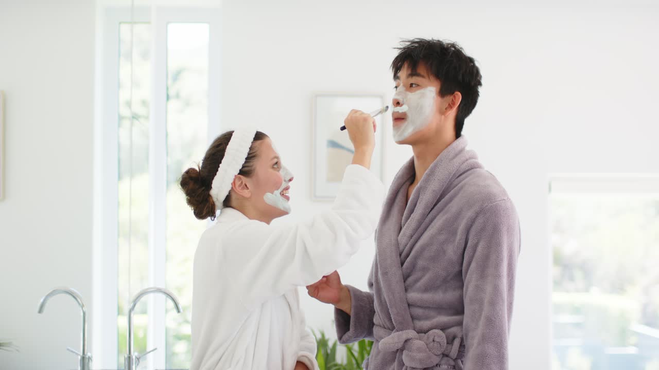 Diverse couple in robes at sink while woman stepping closer applying face mask brush for skincare