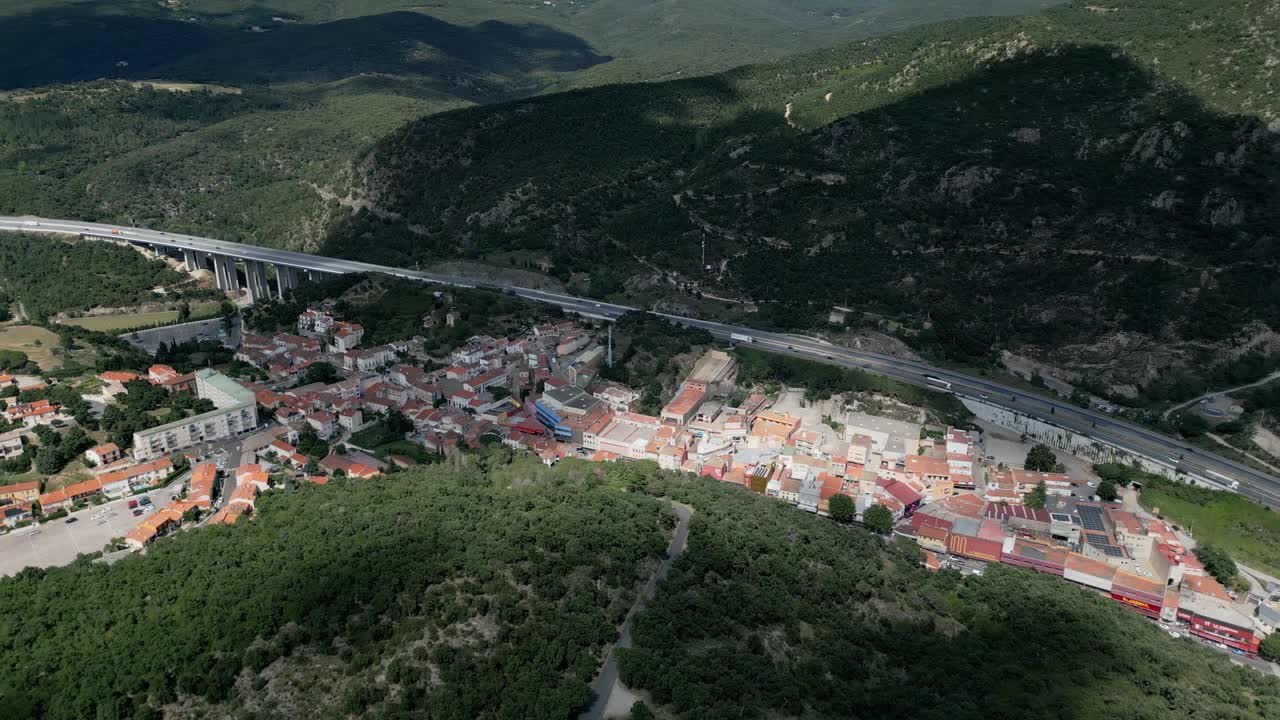 Camera pans over a highway winding through a lush valley in the french pyrenees, revealing the charming village of le perthus nestled amidst the picturesque landscape