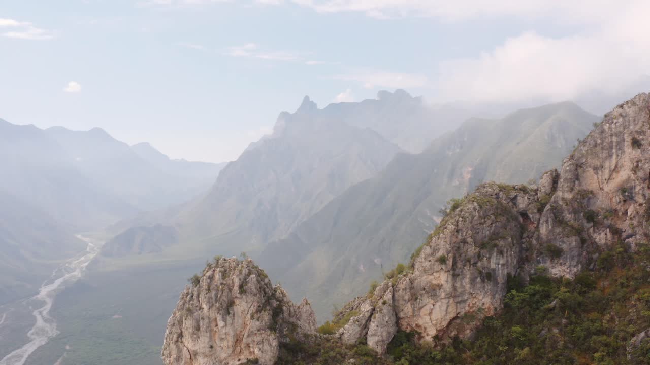 Picos de montanhas escarpados em primeiro plano e céu enevoado com um vale profundo e acidentado e rio ao fundo