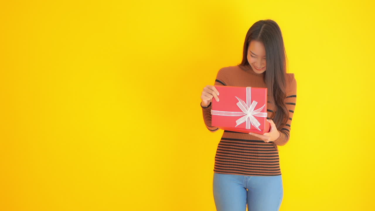 Portrait of a young happy woman having a positive reaction while opening a birthday, anniversary or valentine's day romantic gift on yellow studio background