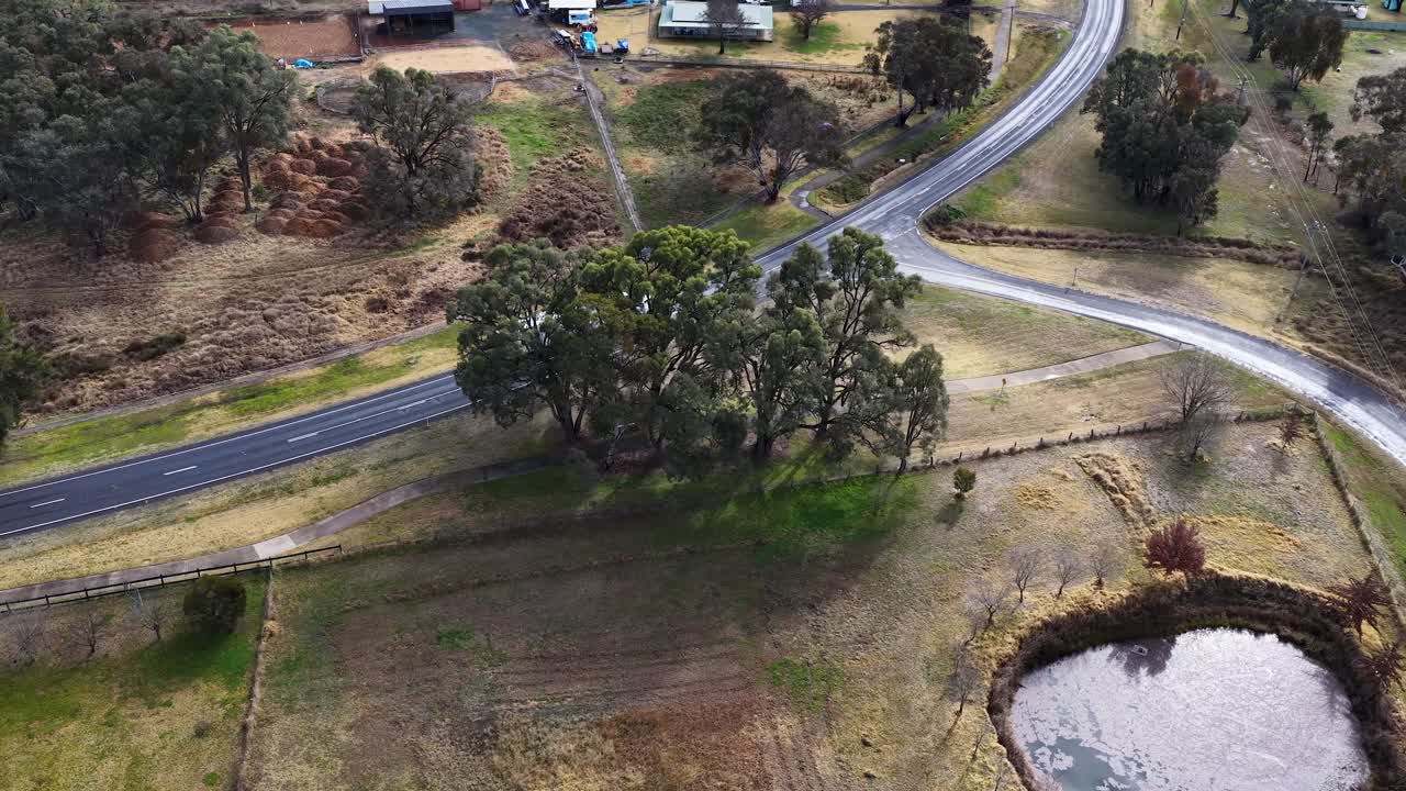 Aerial footage tracks a white semi truck navigating a curved countryside road past rural buildings and ponds in Coonabarabran, Australia, under natural daylight