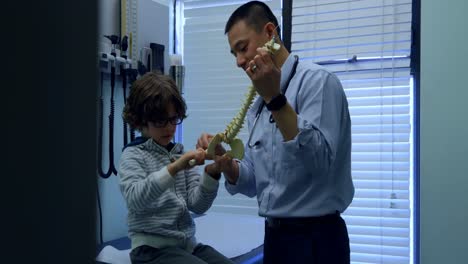 Vista-Lateral-De-Un-Joven-Médico-Asiático-Que-Muestra-Un-Modelo-De-Columna-Vertebral-A-Un-Paciente-Niño-Caucásico-4k