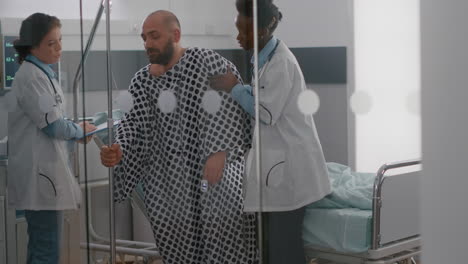 médicos practicantes ayudando al paciente a levantarse de la cama sosteniendo la bolsa de goteo de líquido intravenoso