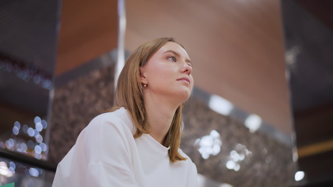 женщина сидит вдумчиво в помещении с спокойным и размышляющим выражением лица, смотря вверх, мягкое освещение подчеркивает ее черты, создавая элегантную и спокойную атмосферу