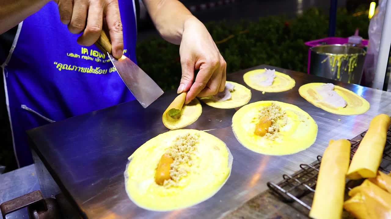 Vendor prepares traditional Thai crispy pancakes at night market. Bright lighting highlights detailed cooking process on a hot griddle