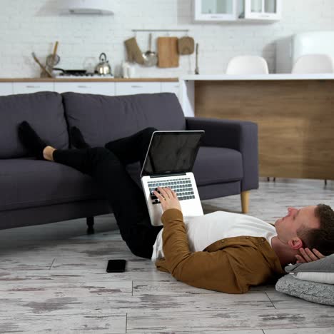 Freelancer working at home. Tired student studying on a laptop while laying on a floor with his legs on sofa. Remote job