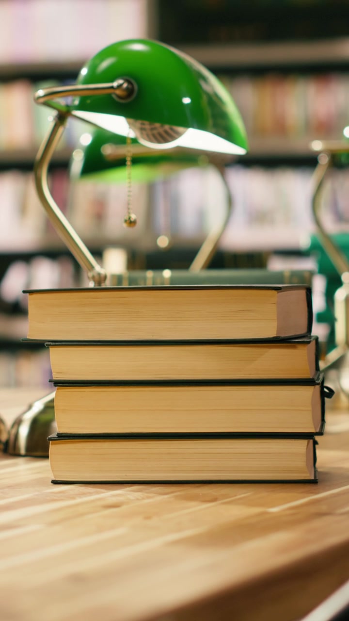 Video verticale. Alta pila di libri su un tavolo della biblioteca con lampade verdi sullo sfondo di scaffali di libri.