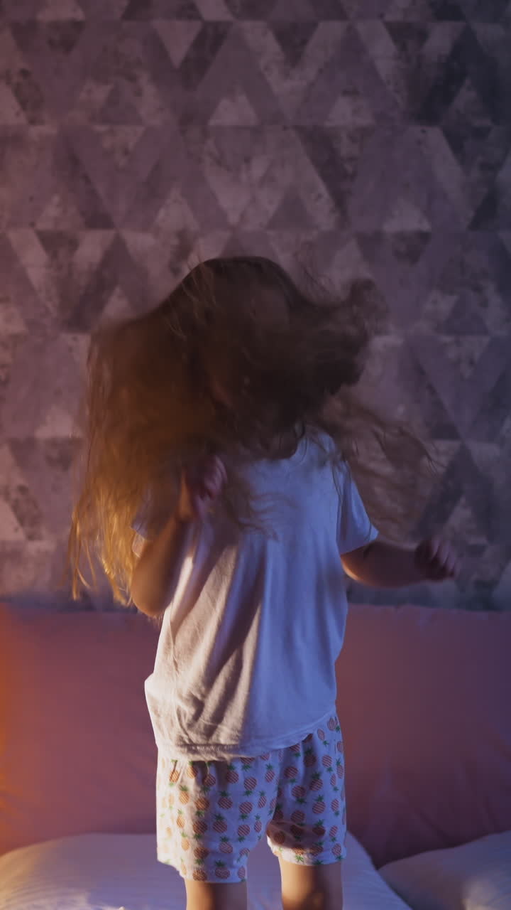 Disobedient little girl with long blonde hair jumps on comfortable bed near play teepee with night lamp inside in dark bedroom in evening slow motion