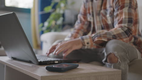 Ventana-Emergente-De-Twitter-Que-Distrae-A-Un-Joven-Profesional-Independiente-Que-Trabaja-Desde-Casa-En-Su-Sala-De-Estar-Usando-Una-Computadora-Portátil,-Distraído-De-Su-Teléfono-Móvil,-Tablas-De-Surf-En-El-Fondo,-Usando-Una-Camisa-A-Cuadros-Naranja