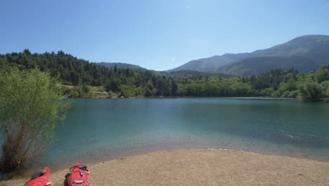 zooming in towards two kayaks next to the clear blue waters of lake tsivlou can be seen as the beautiful greek destination shows tree-covered mountains in green with clear blue skies on the horizon