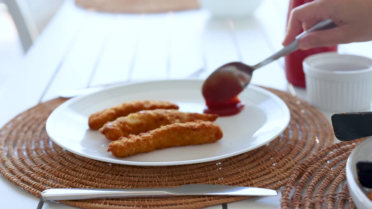 A hand pours ketchup onto fried chicken on a white plate in a well-lit dining area