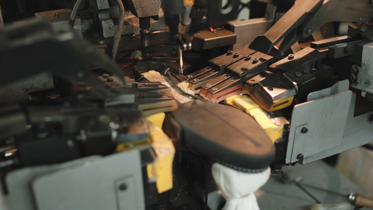 Close-up of a leather shoe sole being shaped and pressed by an industrial lasting machine with metal clamps and yellow components in factory