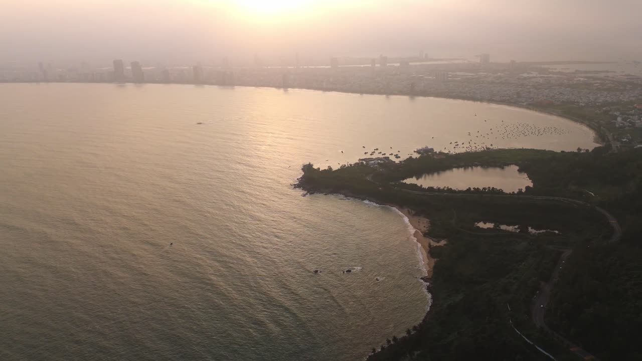 Aerial sunset over the distant Da Nang city skyline and coastline, from the Son Tra Peninsula as city lights begin to glow