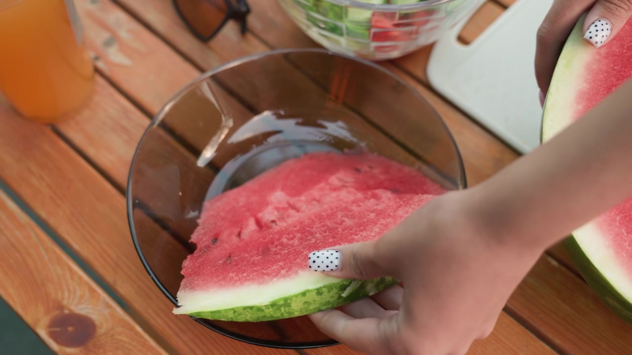 Une femme coupe une grosse pastèque sur une table en bois, plaçant chaque morceau dans des bols en verre transparents. La scène inclut des légumes frais comme des concombres et des tomates en arrière-plan, sous une lumière naturelle.