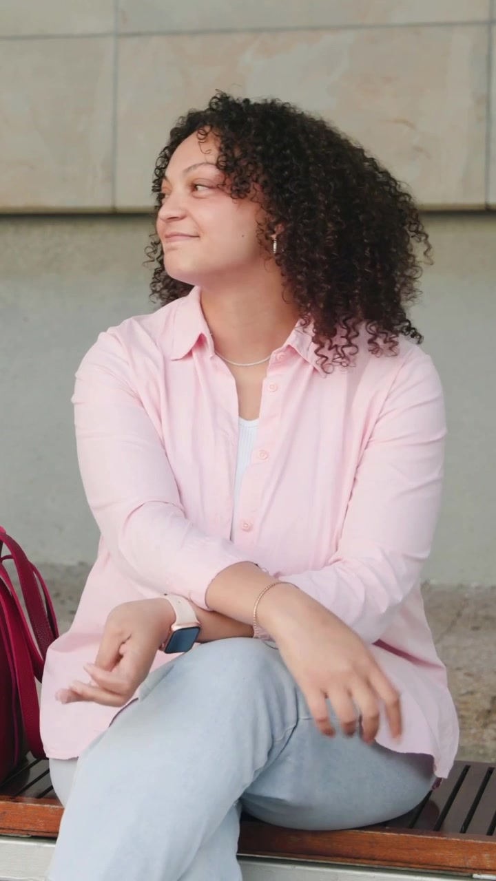 Video verticale: Donna seduta con camicia rosa e jeans chiari che controlla lo smartwatch su una panchina, zaino rosso