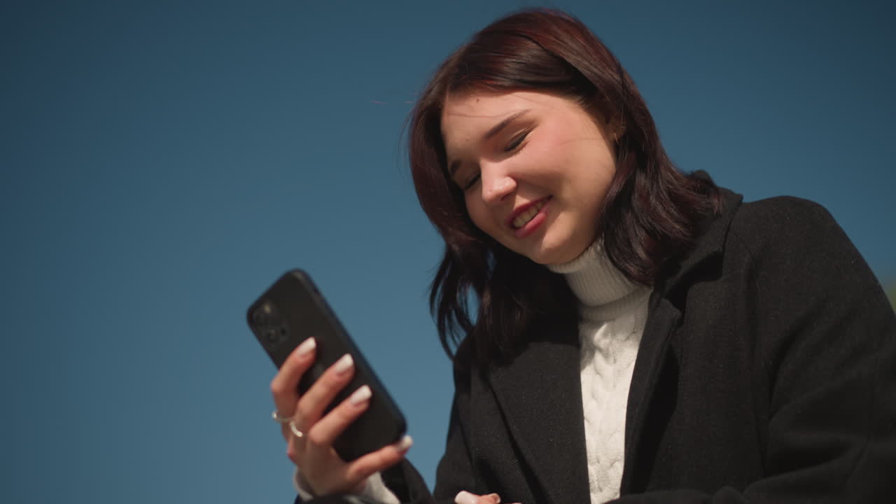 흑색 코트와 색 스웨터를 입은 여성이 밖에서 스마트폰을 들고 있는 클로즈업, 푸른 하늘이 배경으로 작용하면서 그녀는 미묘하게 미소 짓고, 그녀의 손은 반지로 장식되어 우아함을 반영합니다.