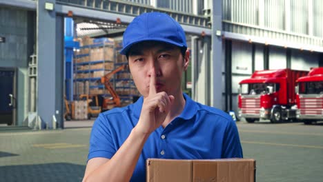 close up of asian male courier in blue uniform showing silence gesture and shaking his head while delivering a carton, outside of logistics distributions warehouse