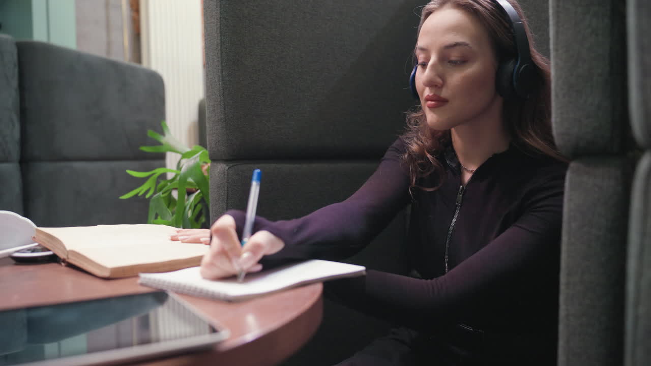 Light skin lady with brown hair wears black headset, writes with one hand in notebook while holding open book with other hand, seated at wooden table in cozy indoor workspace with plant nearby