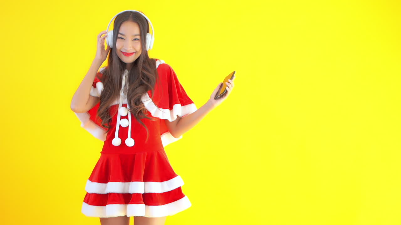 Static shot of a sexy Asian lady wearing a Santa costume and listening to music on her headphones and tablet