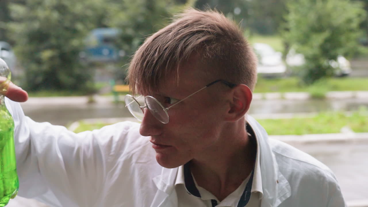 Portrait view of young man in white coat sitting outdoors enjoying snack, chewing while looking aside with relaxed expression during break in urban setting near greenery and wet pavement