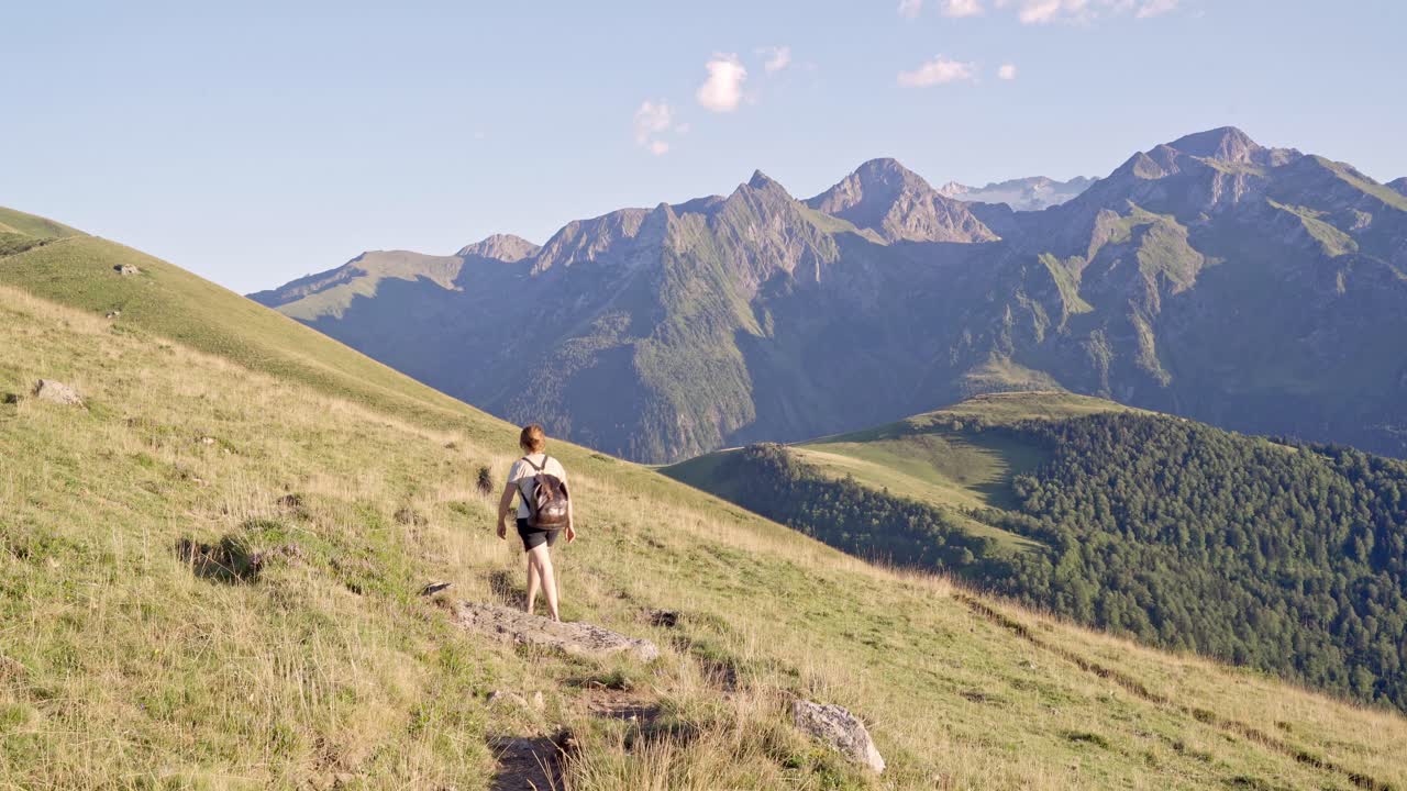 mujer irreconocible caminando por la ladera de la colina de hierba contra las montañas rocosas