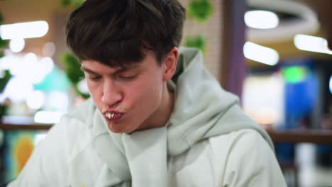 adult tying sweater round neck while gulping burger filled with coleslaw, closeup showing enthusiastic bite, casual interior setting, hand adjusting collar mid bite, messy filling spilling slightly