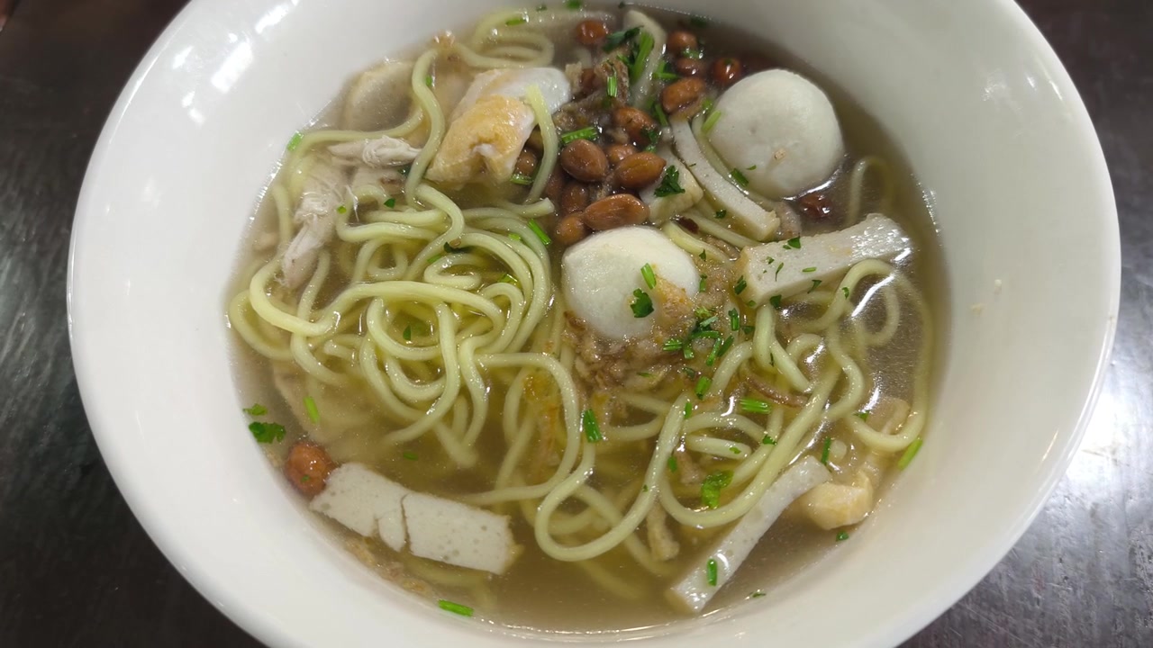 Zuppa di noodle asiatica servita calda in una ciotola di porcellana bianca. Noodle gialli con polpette di pesce, tortini di pesce e verdure miste in un brodo leggero e trasparente. Presentazione del pasto fresca e invitante.