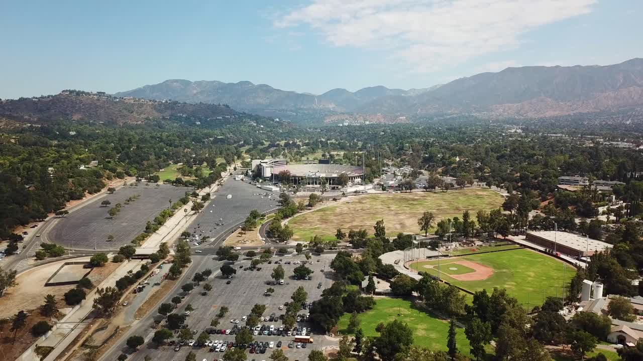 Baseball field, parking cars and rose bowl stadium with mountains in background. Sunny day summer day in Pasadena, California. Drone wide shot